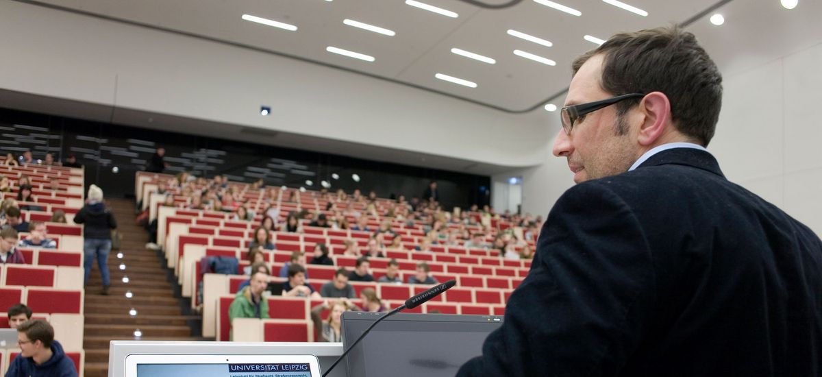 Foto: ein Mann steht am Rednerpult im voll bestetztem Hörsaal und spricht zu den Studierenden während er auch auf einen Laptop blickt.