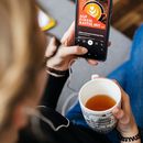 Studentin mit Smartphone und Teetasse. Schulterblick auf ein Smartphone-Display mit geöffnetem Podcast "Auf einem Kaffee mit". 