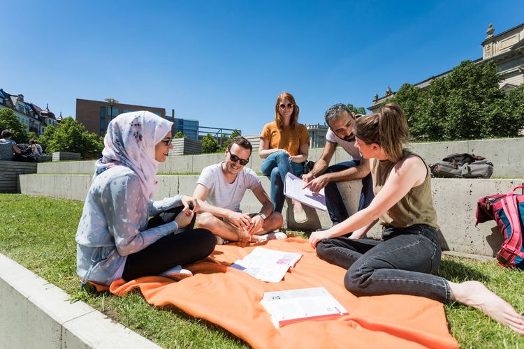 Internationale Studierende sitzen auf einer Picknickdecke in der Sonne vor dem Campus Beethovenstraße 