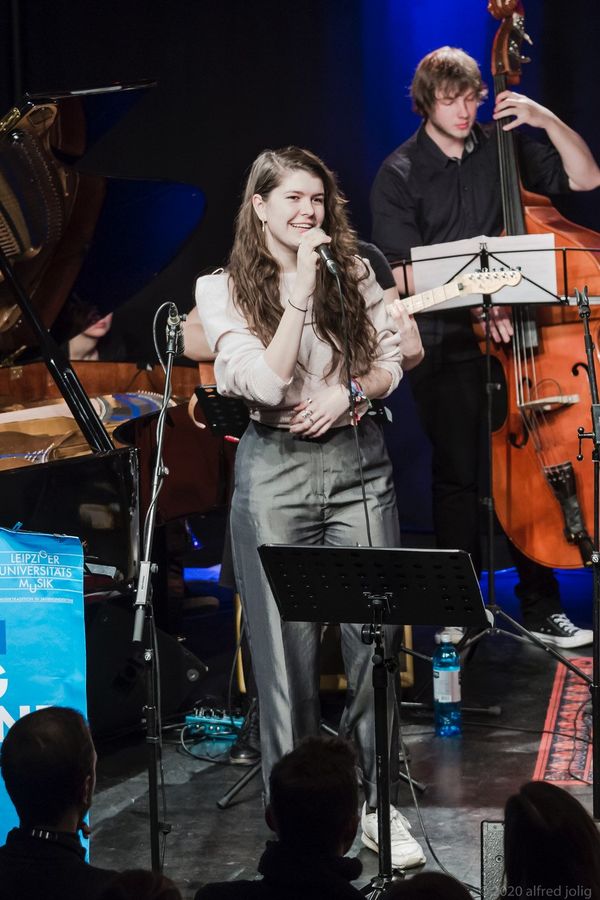 Konzertaufnahme der Unibigband mit einer junge Sängerin im Vordergrund. Im Hintergrund sind Musiker:innen mit einem Kontrabass und einer E-Gitarre zu sehen.