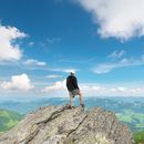 Farbfotografie: Ein Mann steht auf einem Felsen und blickt ins Tal. Der Mann steht mit dem Rücken zur Kamera. Vor ihm liegt ein weites, tiefes, grünes Tal. Am blauen Himmel befinden sich einzelne Wolken.